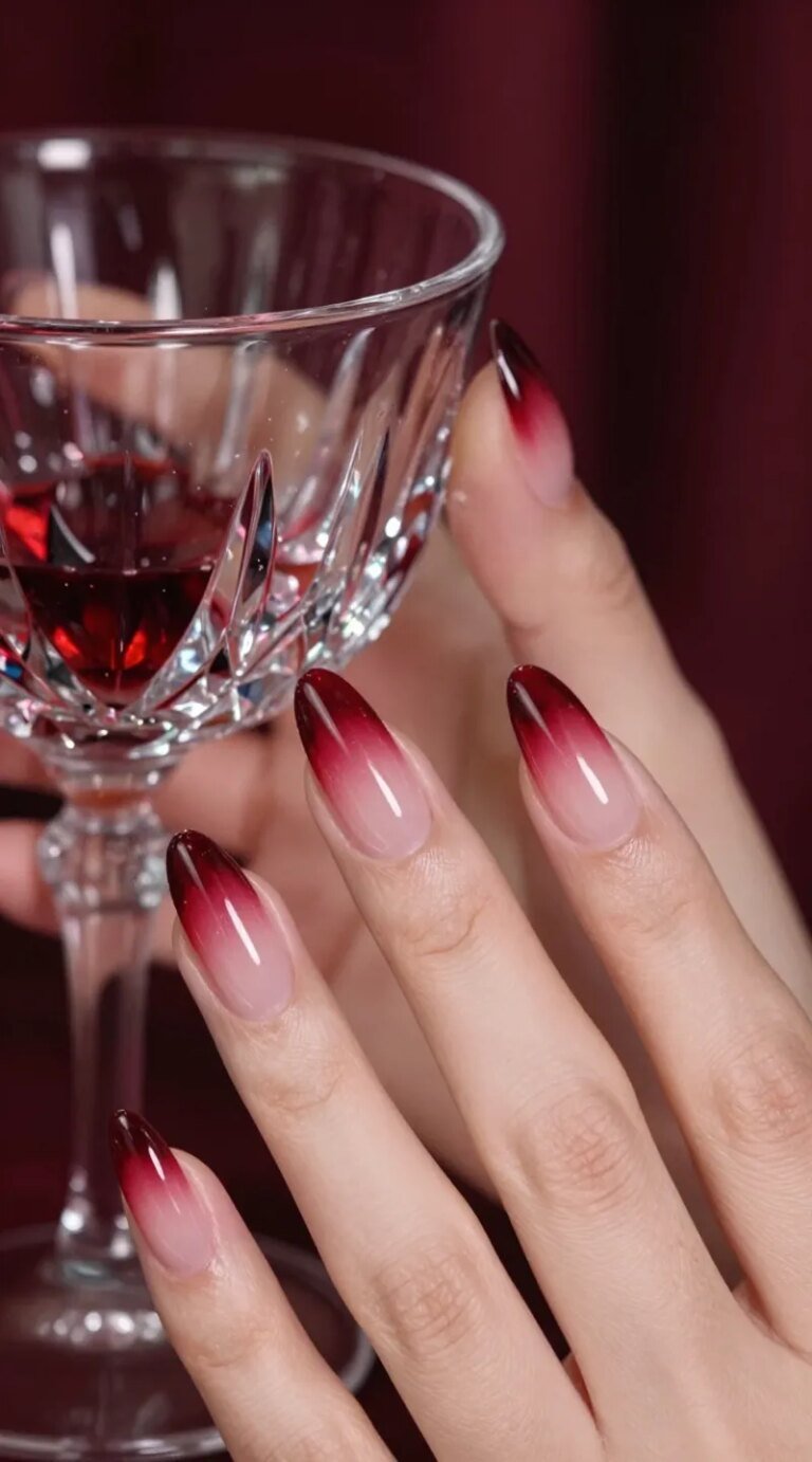 Ultra-realistic close-up beauty photo of a hand with almond-shaped “black cherry glass” tips: sheer jelly cherry-red nails with a translucent, stained-glass effect over a clear-to-soft-pink natural base. The color deepens subtly toward the tips with layered gradient depth, finished with a mirror-like high-gloss top coat. Skin is softly lit, warm undertones, moisturized cuticles, and elegant finger positioning holding a faceted crystal coupe glass with ruby reflections. Background is moody, luxe bokeh in deep burgundy and noir tones for a sultry, expensive vibe—romantic evening ambience. High-resolution macro detail showing the glassy transparency, edge clarity, and wet-shine surface. Natural lighting with gentle specular highlights, shallow depth of field, editorial beauty styling, no heavy props, no logos. Photorealistic, cinematic, intimate, date-night chic.